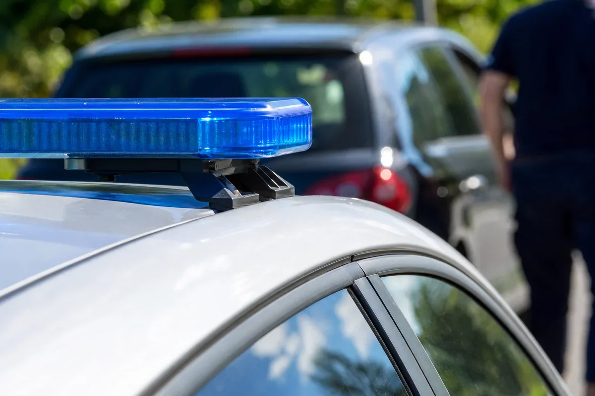Błyskawica policyjnego radiowozu na tle zaparkowanego samochodu i stojącego obok funkcjonariusza.