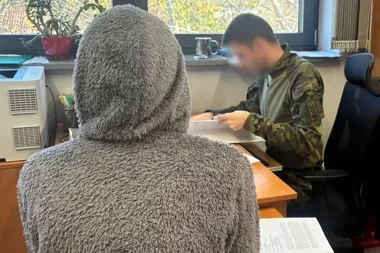 Osoba w szarej, puszystej bluzie z kapturem siedzi plecami do obiektywu przy biurku, naprzeciwko żołnierza w mundurze, który przegląda dokumenty w pomieszczeniu