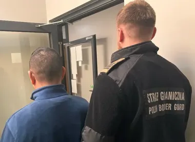 Funkcjonariusz Straży Granicznej prowadzi mężczyznę w niebieskiej bluzie przez korytarz z drzwiami szklanymi.