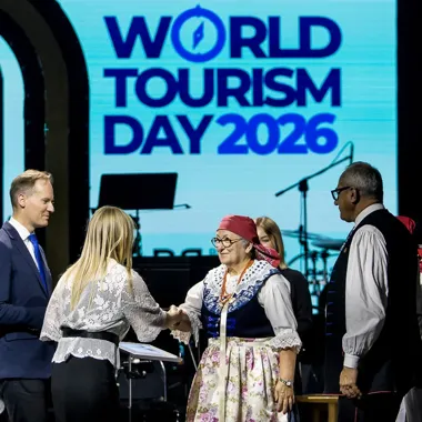Uczestnicy oficjalnej uroczystości z okazji Światowego Dnia Turystyki 2026 witają się na scenie przed ekranem z hasłem wydarzenia.