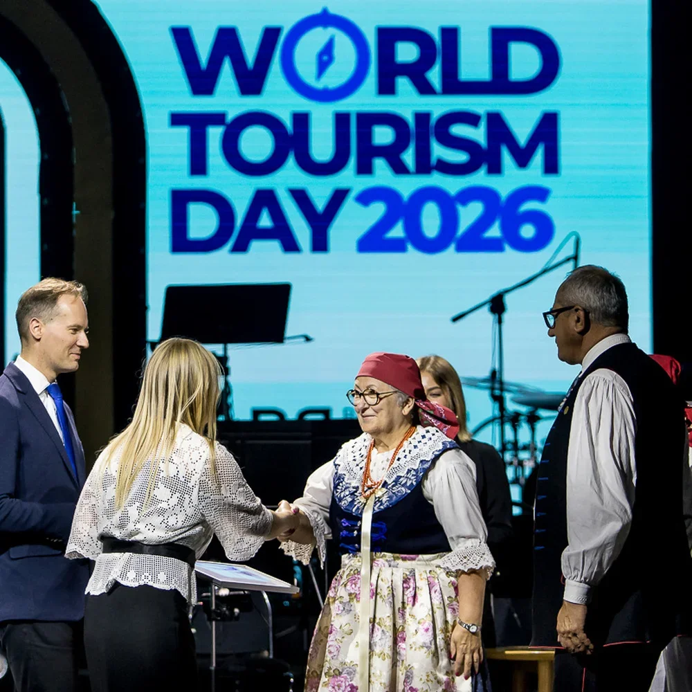 Uczestnicy oficjalnej uroczystości z okazji Światowego Dnia Turystyki 2026 witają się na scenie przed ekranem z hasłem wydarzenia.